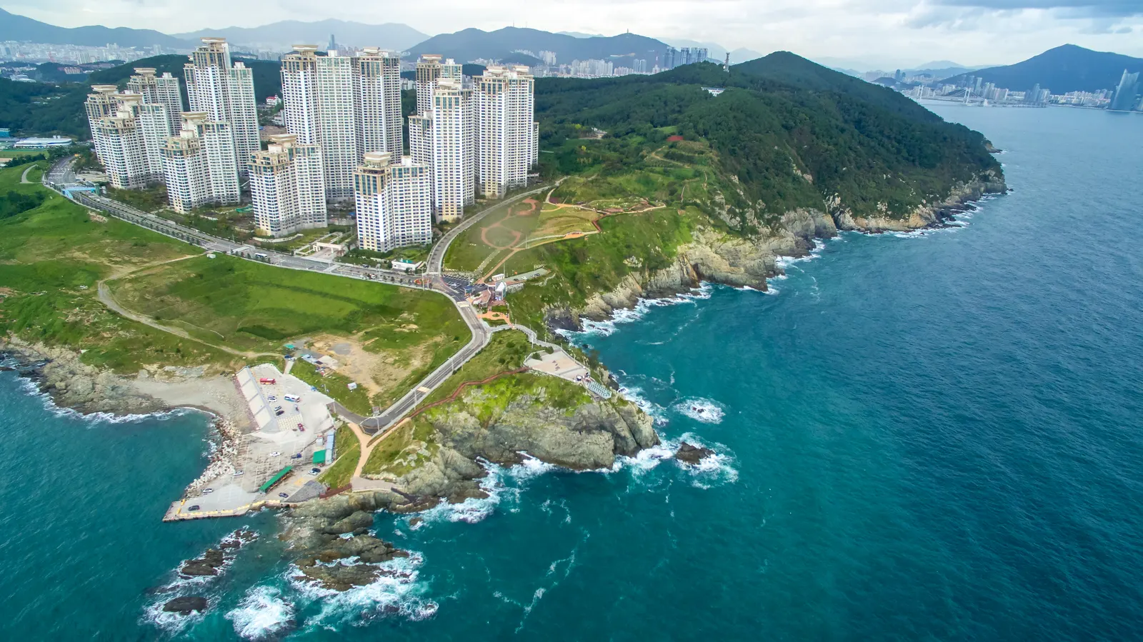 Vista aérea da costa junto a Oryukdo: promontório rochoso, mar azul, encosta arborizada e torres residenciais.